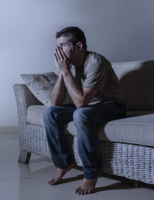 lifestyle dramatic light portrait of young sad and depressed man sitting at shady home couch in pain and depression feeling stressed and desperate crying alone suffering anxiety crisis