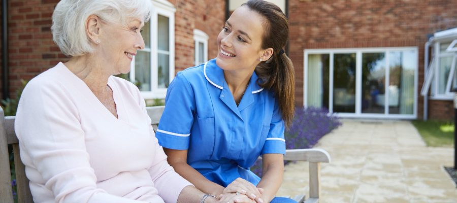 Senior Woman Sitting On Bench And Talking With Nurse In Retirement Home