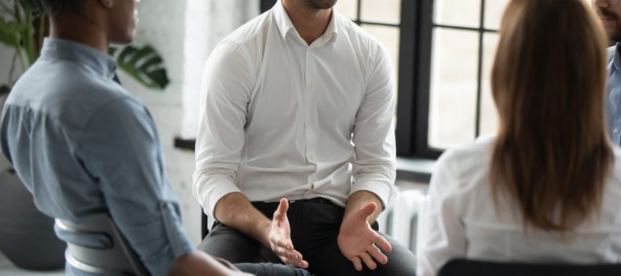 Smiling confident psychologist speaking at group therapy session, young man sharing problems with diverse people sitting in circle, listening to coach at meeting, mentor training staff
